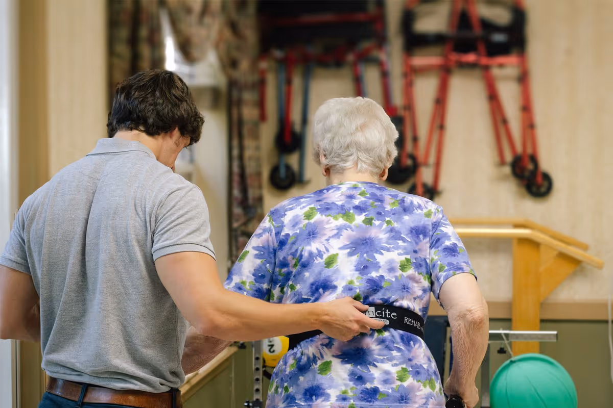 A caregiver assists an elderly woman wearing a floral shirt with a rehabilitation belt in a therapy or exercise room equipped with parallel bars and mobility aids hanging on the wall.