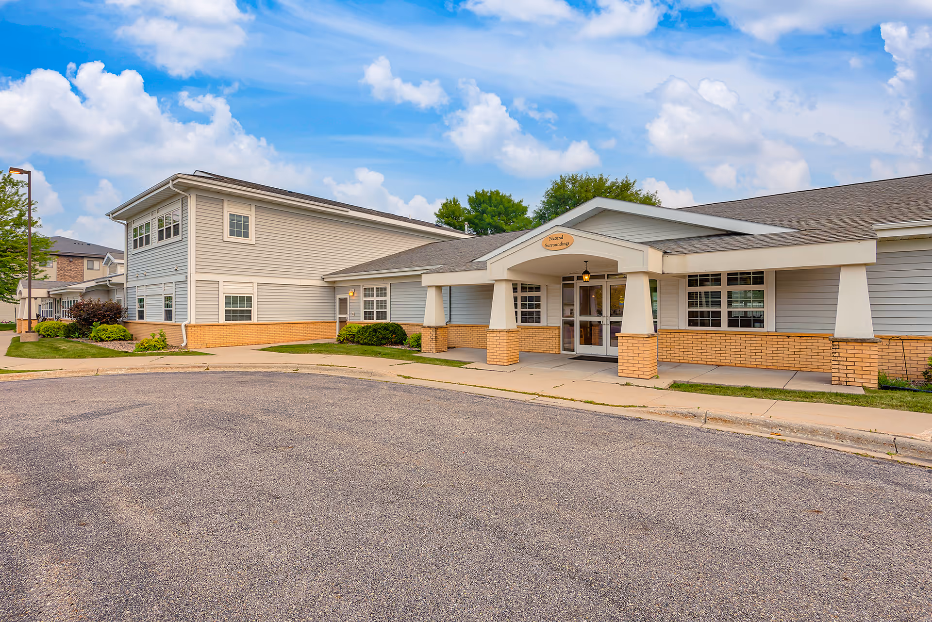 Front exterior of a low-rise senior living building with a covered main entrance, brick base, gray siding, and a circular driveway.