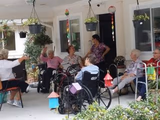 A group of elderly people, some in wheelchairs, sitting and socializing on a covered patio outside a building. There are hanging plants and decorations around the patio area.