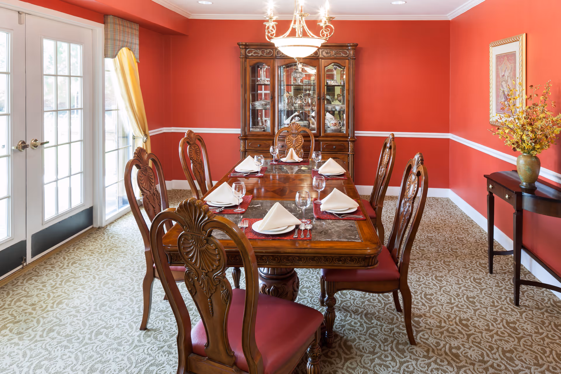 A formal dining room with red walls and white trim, featuring a wooden dining table set for six with folded white napkins, glasses, and silverware. The room has ornate wooden chairs with red cushions, a chandelier overhead, a glass-fronted china cabinet against the back wall, and a side table with a vase of yellow flowers. French doors with yellow curtains allow natural light into the room.