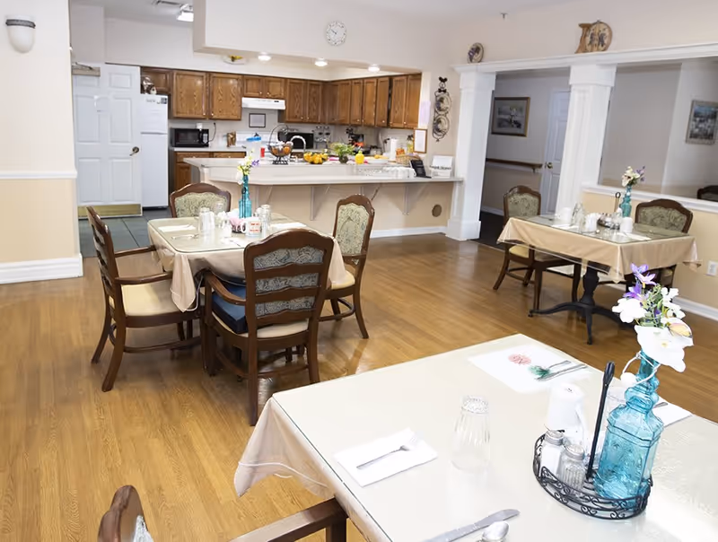 A dining area in a senior living facility with several tables covered with beige tablecloths, each set with utensils, glasses, and small flower vases. In the background, there is a kitchen with wooden cabinets, a refrigerator, microwave, and stove. The floor is wooden, and the walls are light-colored.