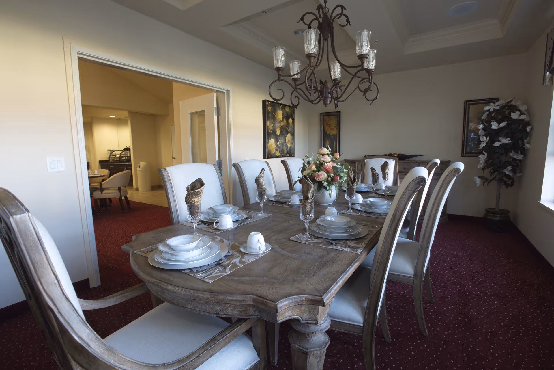 A formal dining room with a wooden table set for eight, upholstered chairs, a floral centerpiece and a chandelier.
