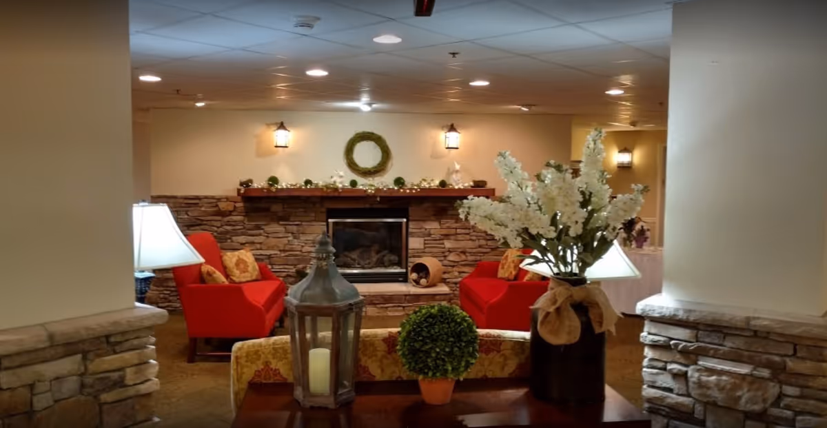 Cozy interior lounge area with two red armchairs facing a stone fireplace decorated with a wreath and string lights. A wooden table in the foreground holds a large lantern with a candle, a small potted plant, and a vase with white flowers. Warm lighting and stone accents create a welcoming atmosphere.