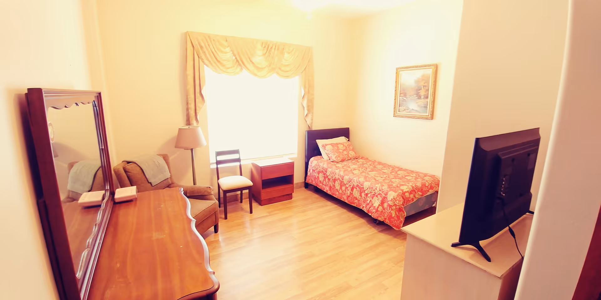 A small, cozy bedroom with a single bed covered in a red floral bedspread, a wooden dresser with a large mirror, a cushioned armchair, a wooden chair, a small wooden nightstand, a floor lamp, and a flat-screen TV on a stand. The room has light-colored walls, a window with a valance, and a framed picture on the wall.