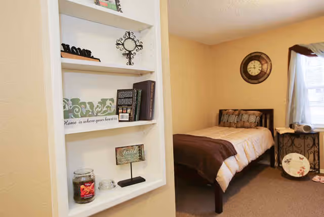 Cozy single-occupancy bedroom with a twin bed, wall clock, window, and a recessed shelf displaying decorative items.
