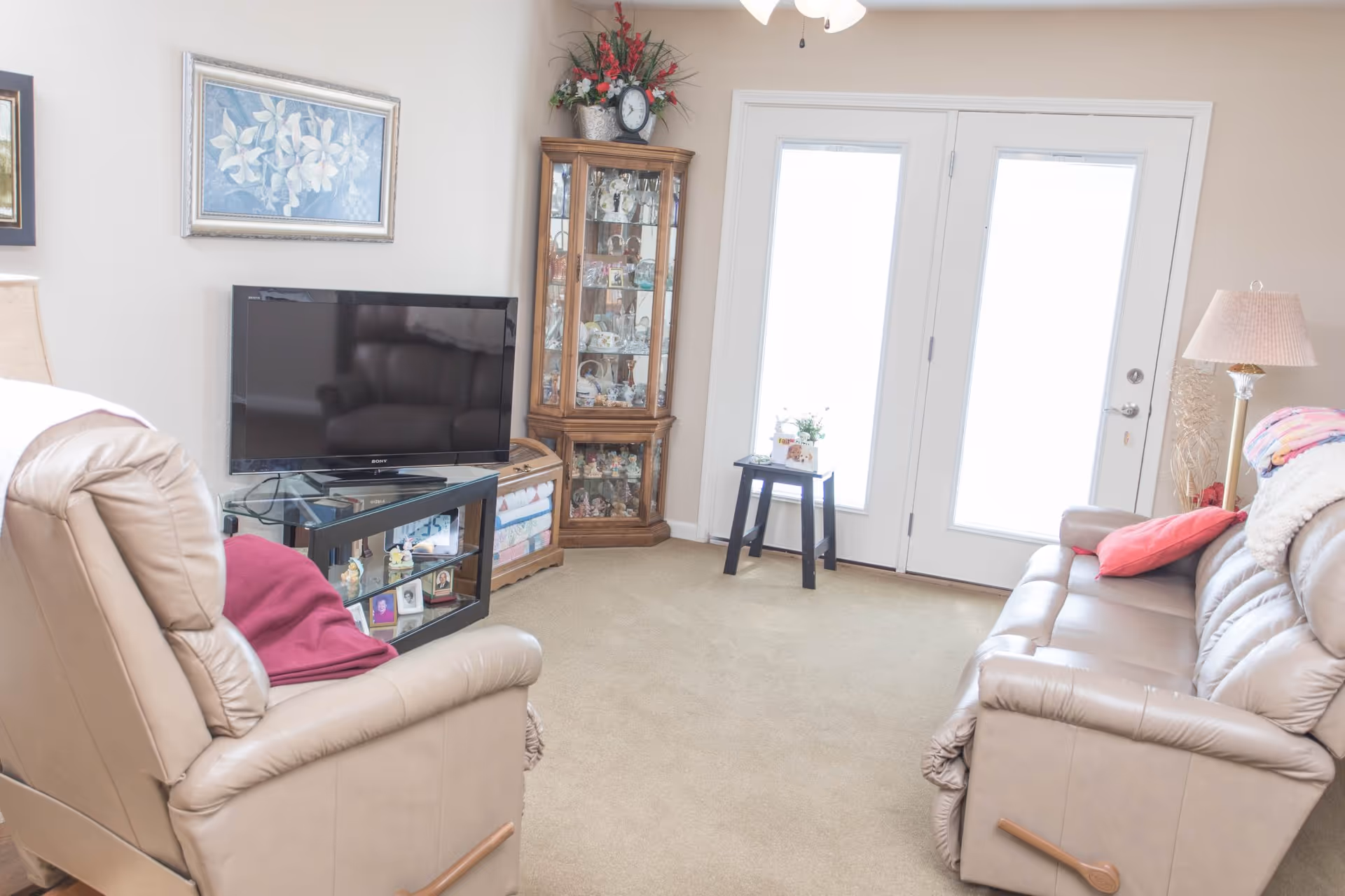 A cozy living room with beige leather recliner and sofa, a glass TV stand holding a flat-screen television, a wooden corner cabinet displaying decorative items, a small black side table with a flower arrangement, and double glass doors letting in natural light.