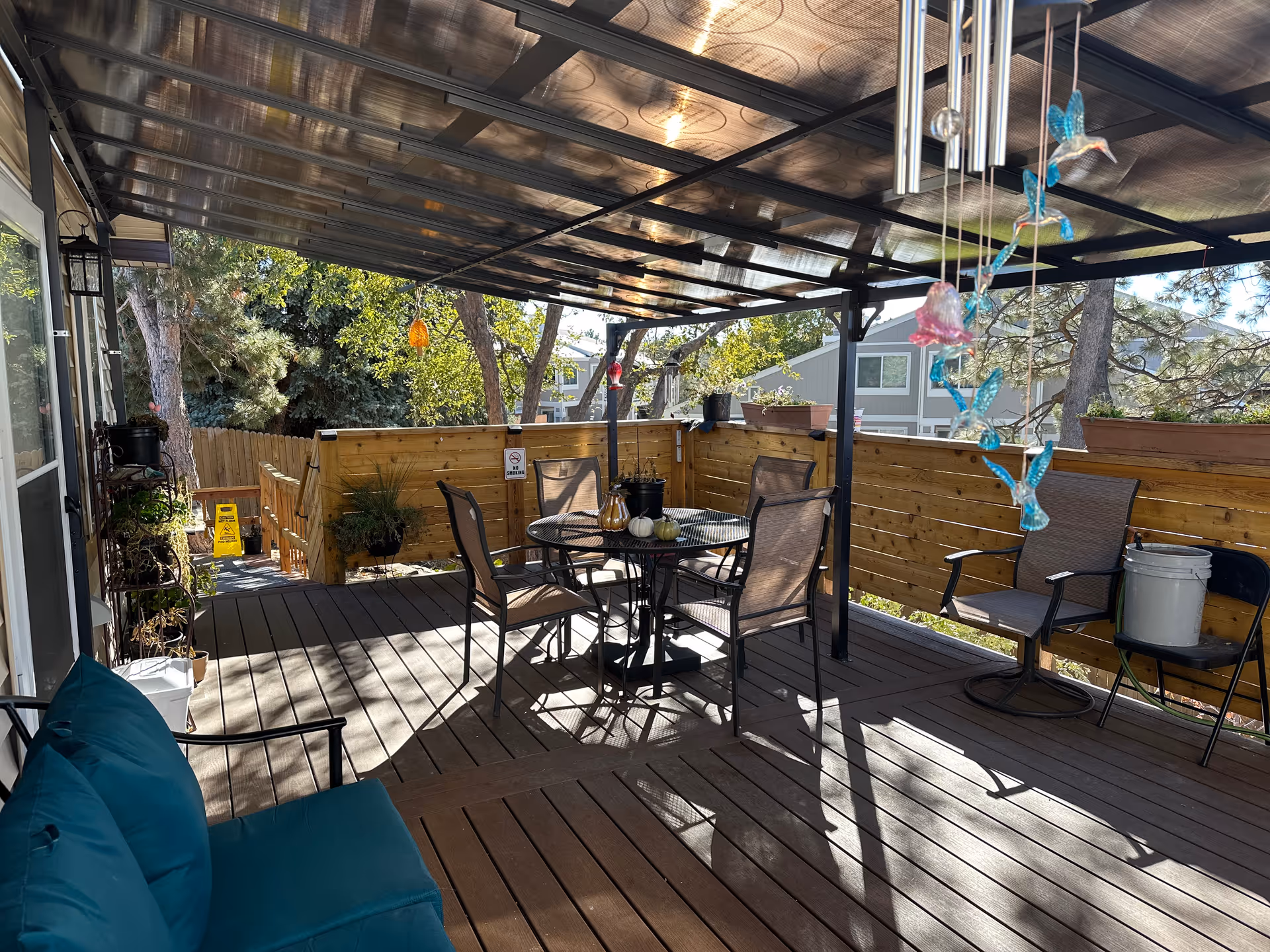 Covered outdoor patio area with a round metal table and four chairs, a cushioned bench, hanging decorative glass birds, potted plants, and wooden fencing surrounding the space. Trees and neighboring houses are visible in the background.