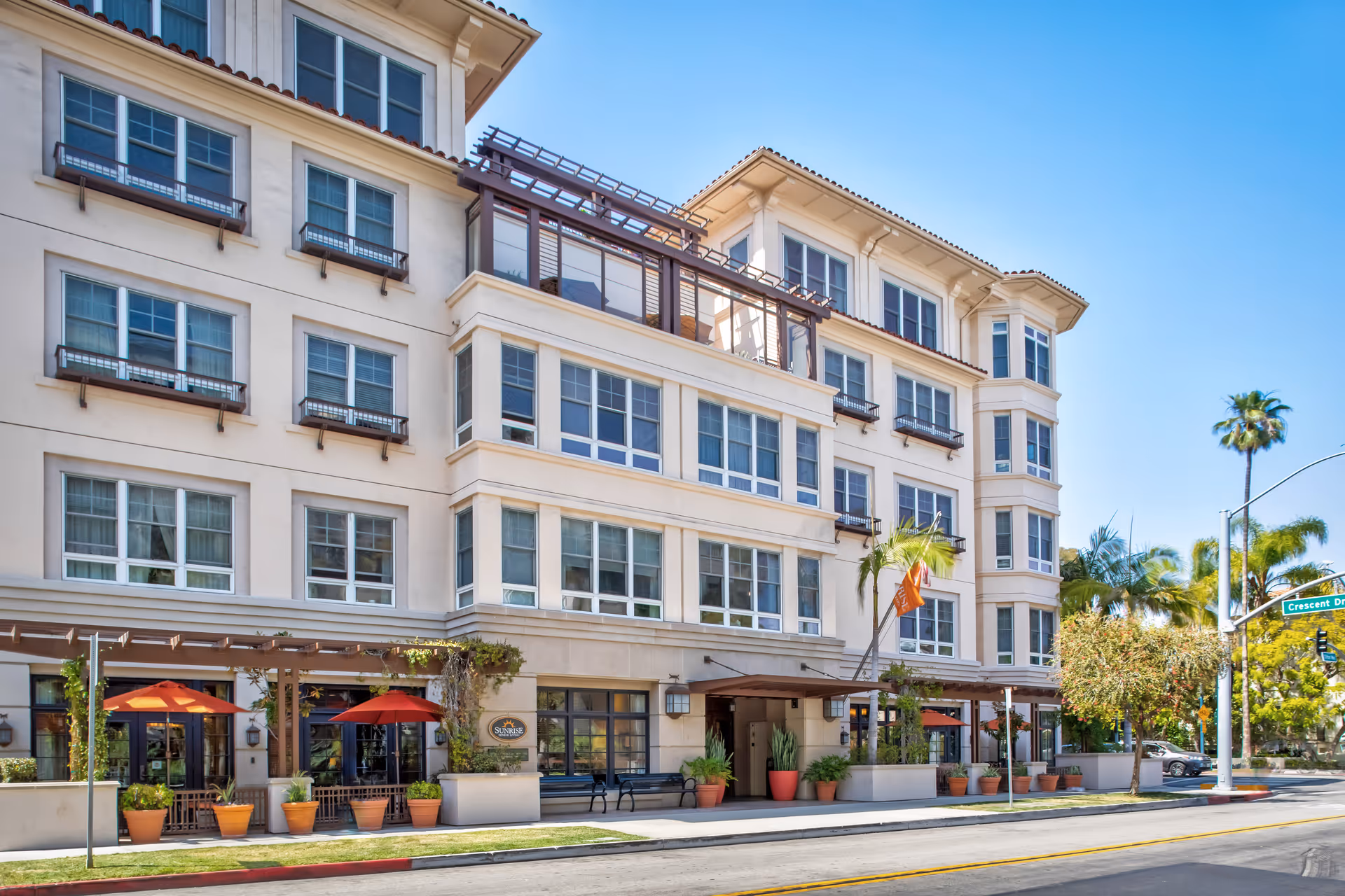 Exterior view of a multi-story senior living facility building with large windows, potted plants, and orange umbrellas shading outdoor seating areas. The building has a beige facade with a tiled roof and a sign that reads 'Sunrise'. A street with a palm tree and a street sign for Crescent Drive is visible in the foreground under a clear blue sky.