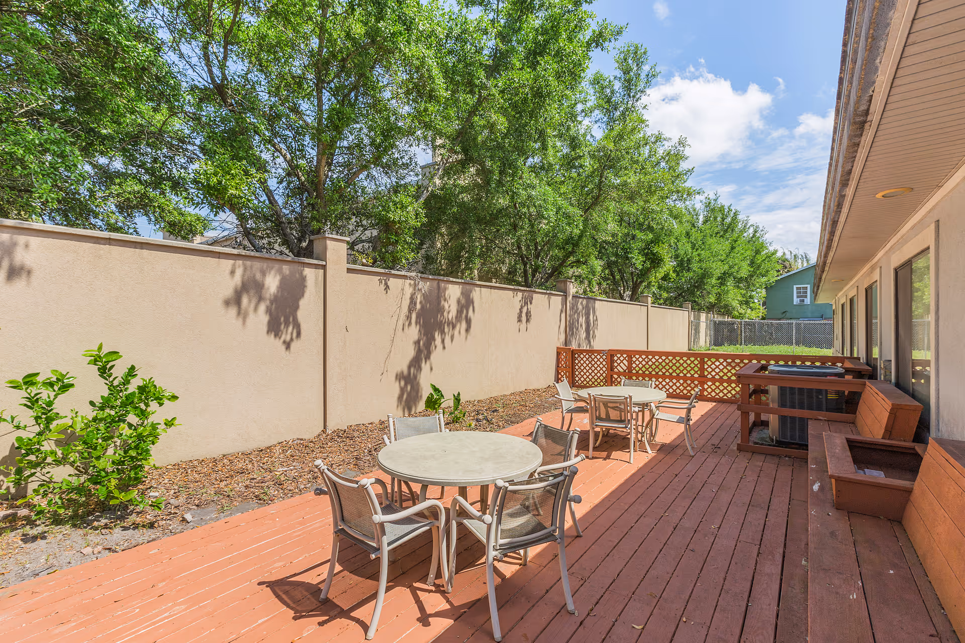 Outdoor patio area with round tables and chairs on a wooden deck next to a building. There is a tall beige wall with trees behind it and a partly cloudy sky above.