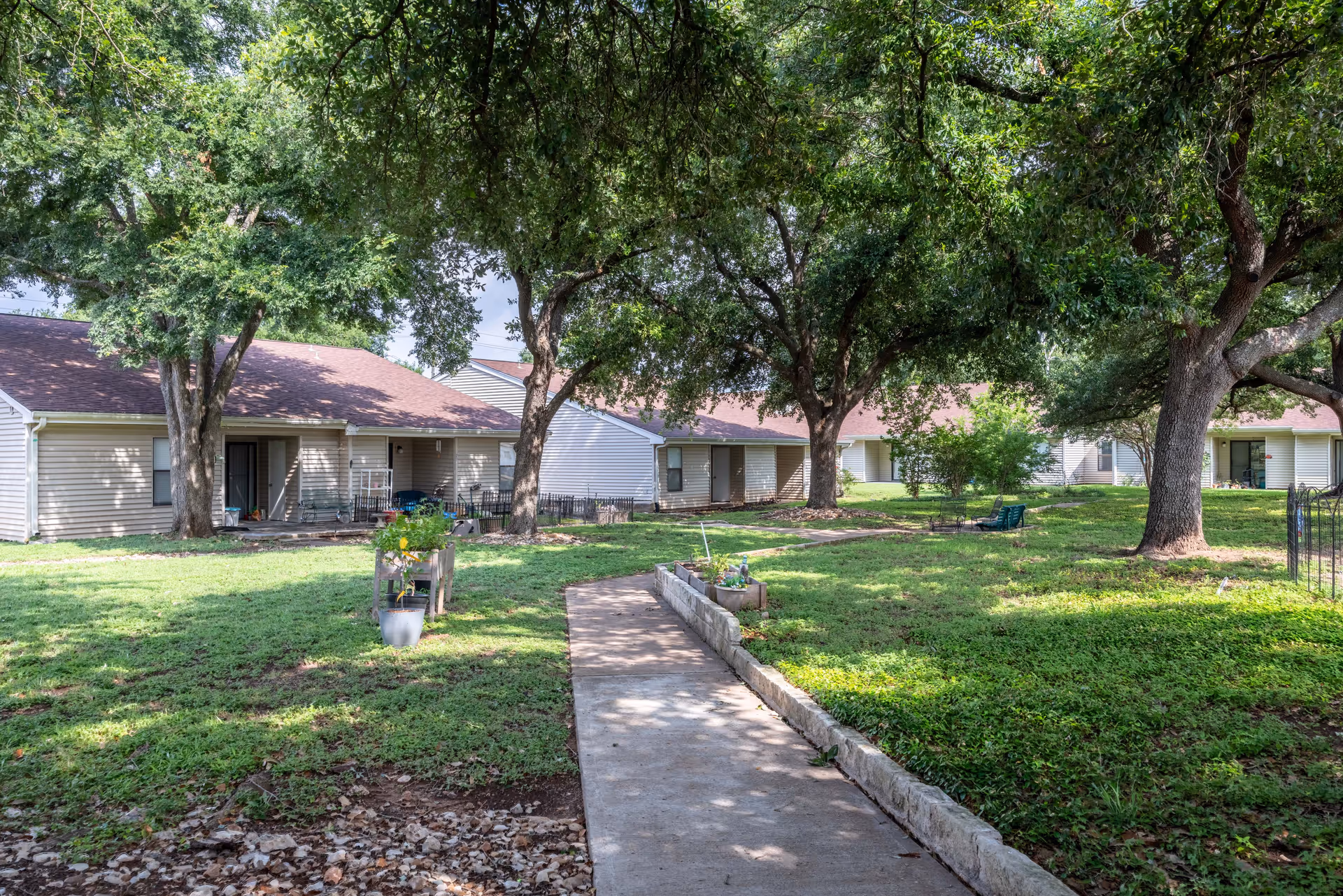 A peaceful outdoor courtyard area at The Clairmont Retirement Community featuring a concrete walkway bordered by a low stone wall, green grass, large mature trees providing shade, and single-story residential buildings with beige siding and brown roofs in the background.