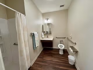 A clean and spacious bathroom with a white toilet, a small sink with a mirror above it, a towel rack with towels, a shower area with a curtain, and a trash bin on the wooden floor.