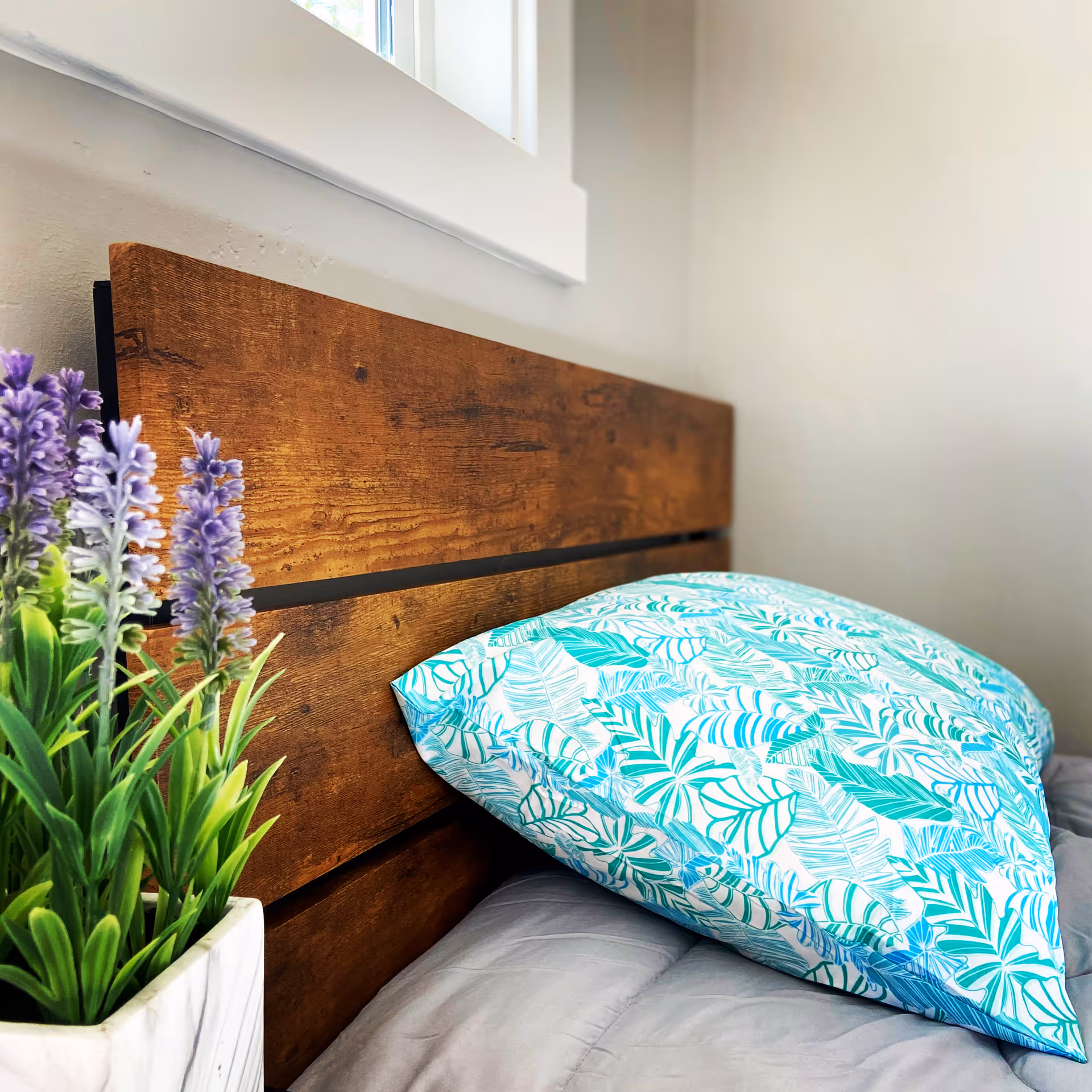 Close-up view of a wooden headboard with a blue and white patterned pillow resting against it, next to a white pot containing green and purple artificial flowers, with a window above.
