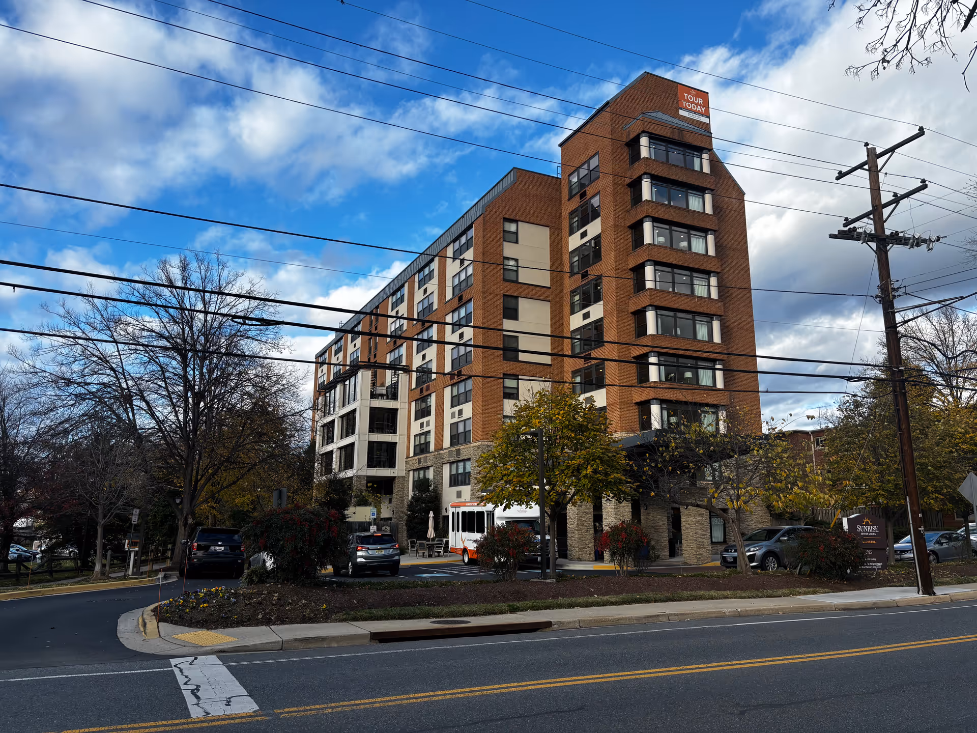 Front exterior of a multi-story brick senior living building with parked cars, trees, and overhead power lines.