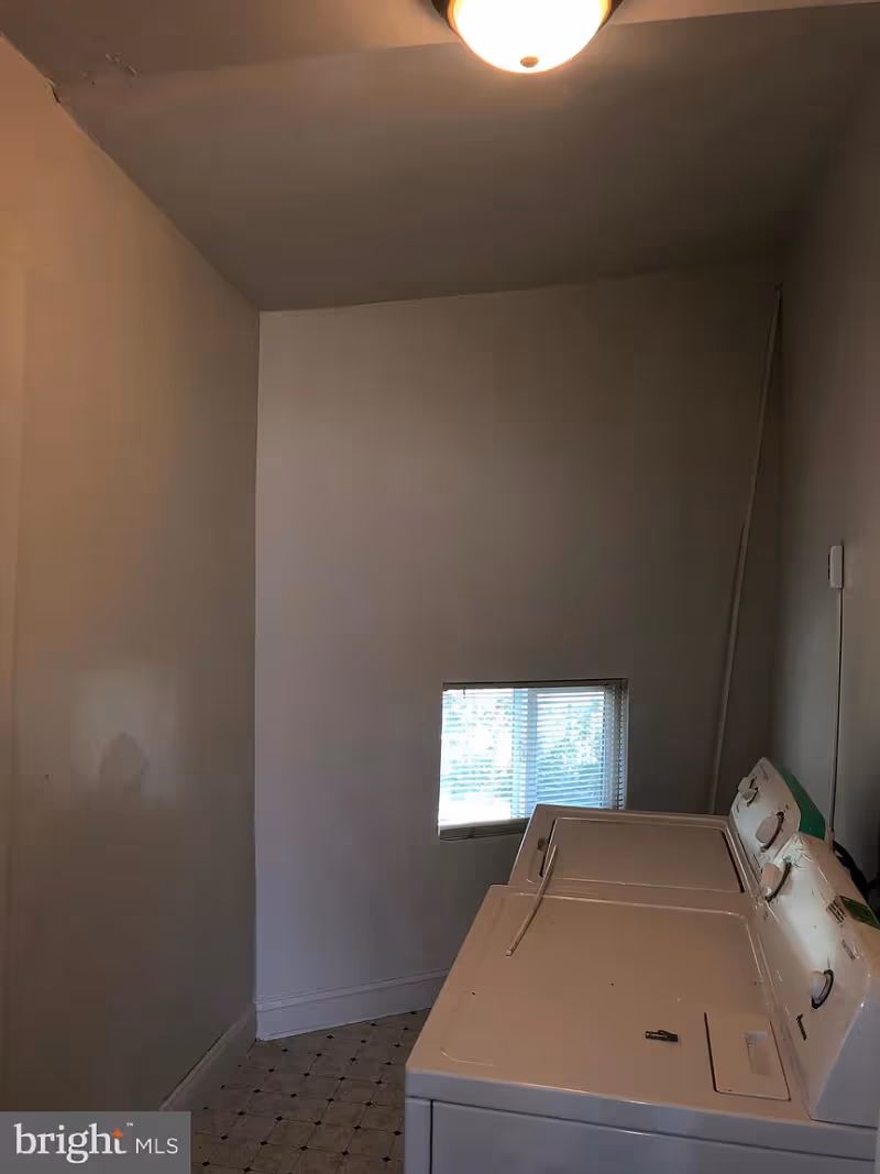 A small laundry room with a white washing machine and dryer side by side against the right wall. There is a small rectangular window with blinds on the back wall and a ceiling light fixture above. The walls are plain and painted white, and the floor has a patterned linoleum covering.