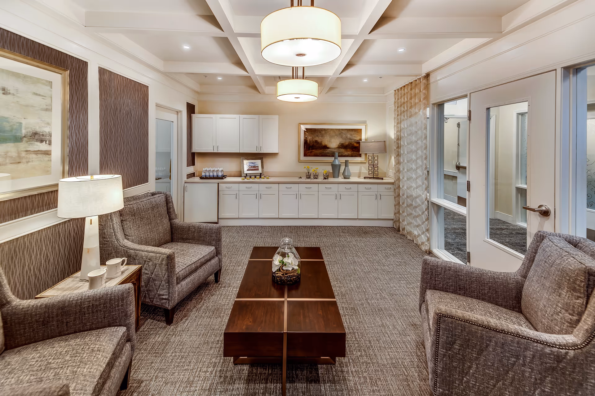 A cozy sitting area in a senior living facility with three gray upholstered armchairs arranged around a dark wooden coffee table. The room features a carpeted floor, a side table with a lamp and two cups, and a long white cabinet with a mini fridge and decorative items including vases and framed pictures. The walls are decorated with framed artwork and patterned wallpaper, and there are two large ceiling lights providing warm illumination.