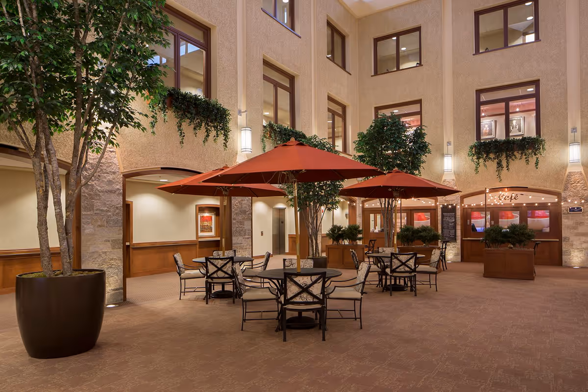 Indoor seating area with round tables and cushioned chairs under large red umbrellas, surrounded by potted trees and plants. The space has beige walls with multiple windows, wall-mounted lights, and a café entrance in the background.