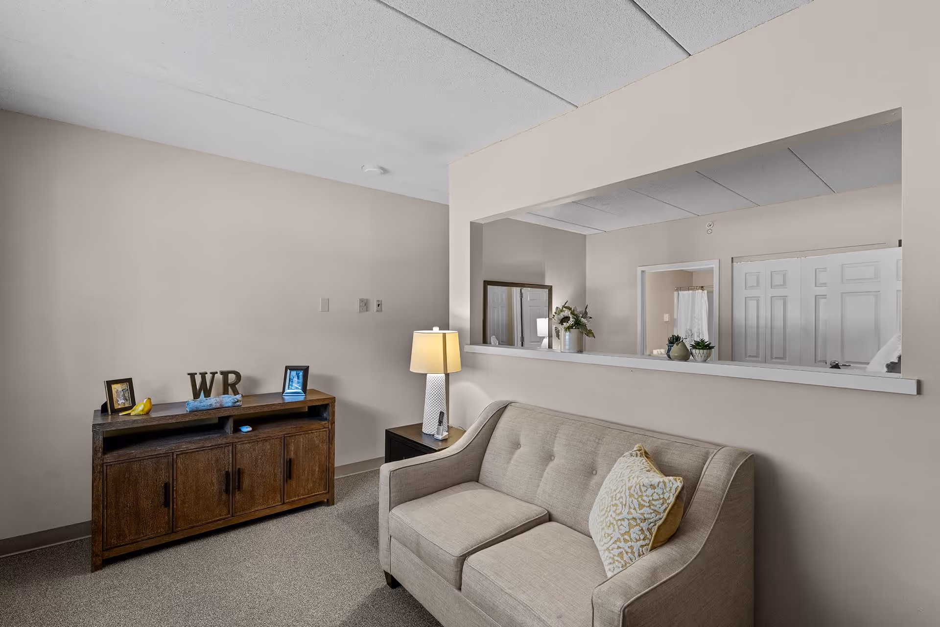 A cozy living room area with a beige sofa adorned with a decorative pillow, a wooden cabinet with framed photos and decorative items on top, a side table with a lamp, and a large interior window opening to another room with white double doors and a mirror.