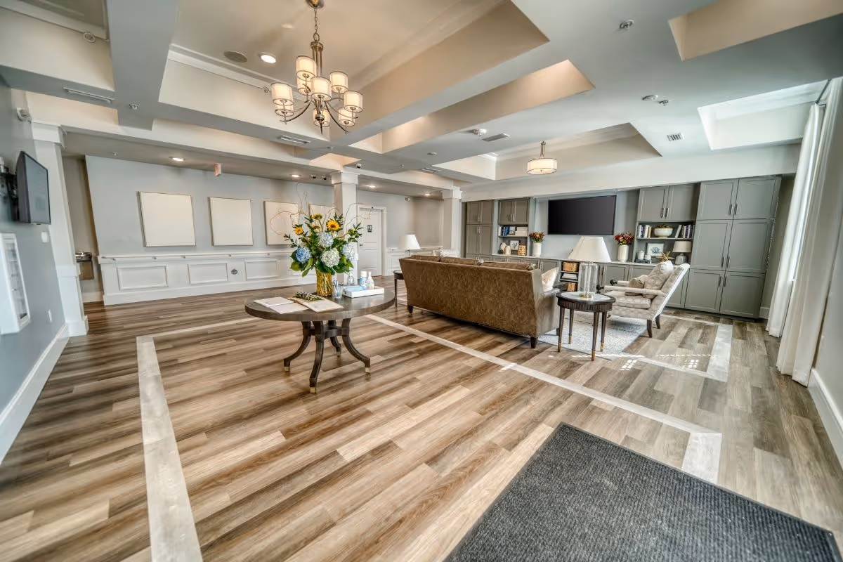 A spacious and well-lit living room area with wood flooring and a central round table holding a vase of flowers. The room features a brown sofa, an armchair, a large flat-screen TV mounted on a wall with built-in cabinets and shelves, and ceiling light fixtures. Large windows with sheer curtains allow natural light to fill the space.
