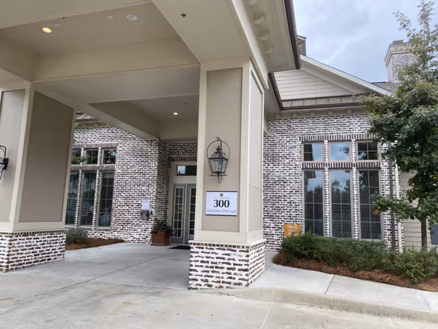 Entrance area of The Claiborne at Hattiesburg Independent Living facility showing a covered drop-off zone with brick and beige pillars, large windows with blinds, a lantern-style light fixture, and a sign displaying the address 300 Pinehurst Pines Blvd.