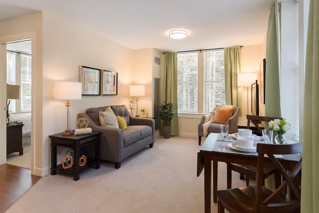 Bright furnished living room with a sofa, armchair, small dining table, and large windows with green curtains.