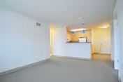 Empty living room with beige carpet looking toward a small open kitchen with a breakfast bar and pendant lights.