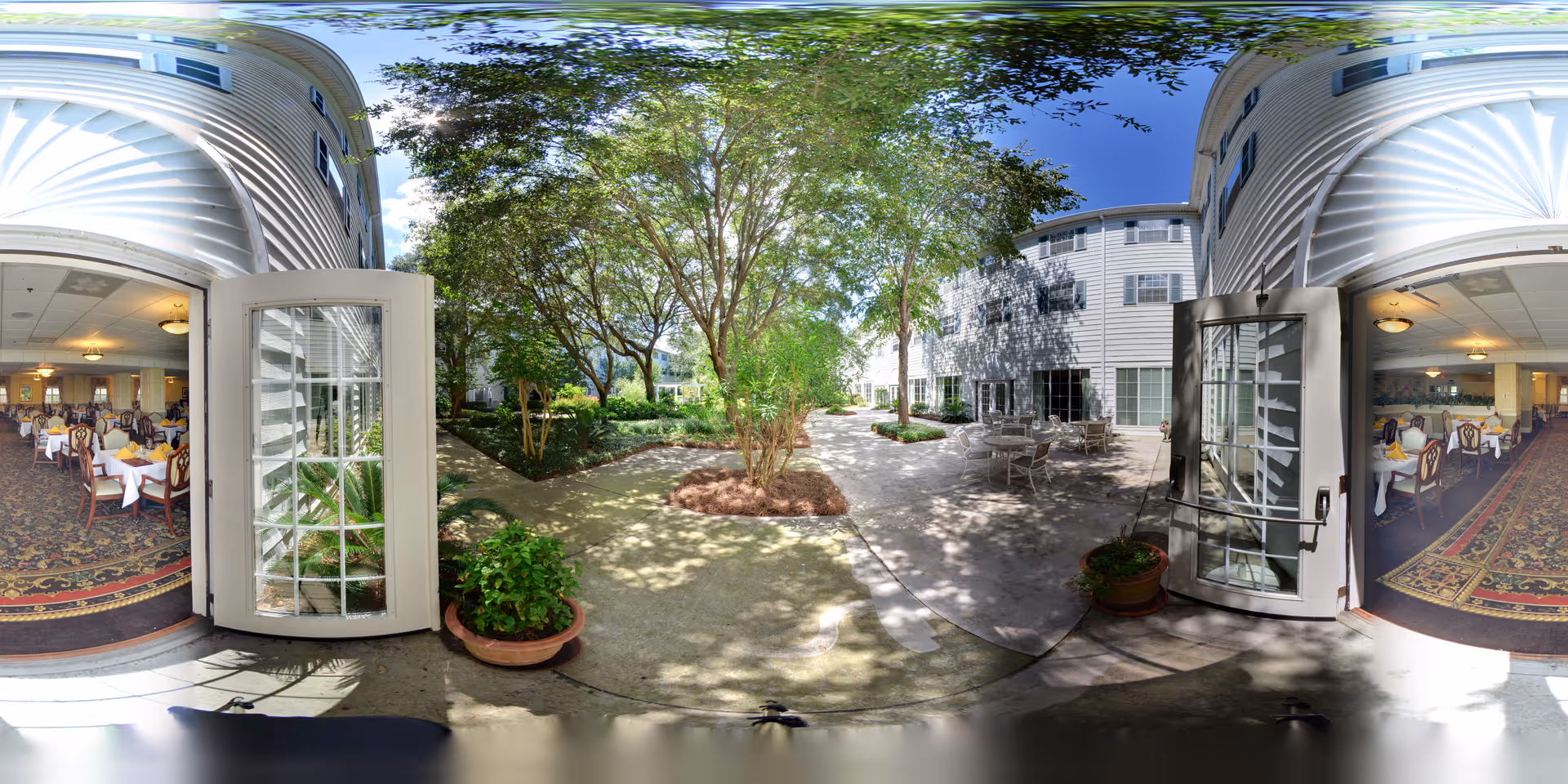 A bright outdoor courtyard area with trees, plants, and seating surrounded by a multi-story white building. Two open glass doors lead from the courtyard into a dining room with tables covered in white tablecloths and chairs arranged neatly. The sky is clear and blue.