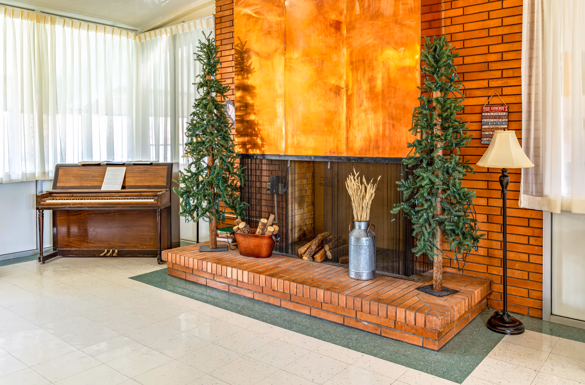 A bright common room featuring a large brick fireplace with decorative trees and logs, a piano by the window, and a floor lamp.