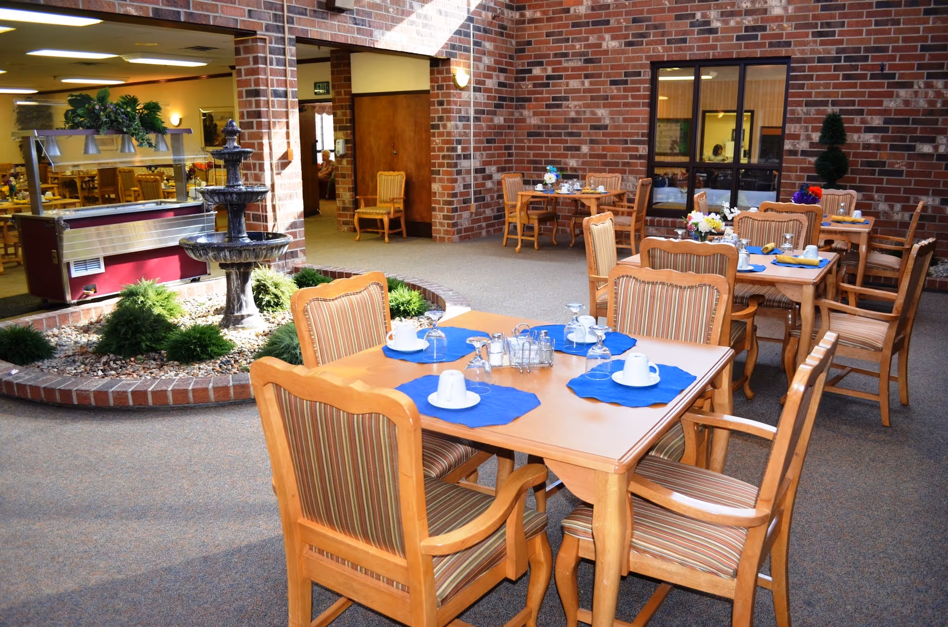 Indoor dining area with wooden tables and chairs set with blue placemats around a central fountain and brick-walled atrium.