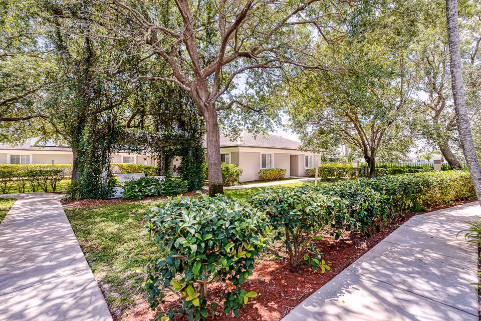 Outdoor garden area at Cottages at Palm Beach with paved walkways, green bushes, large trees providing shade, and a light-colored single-story building in the background.