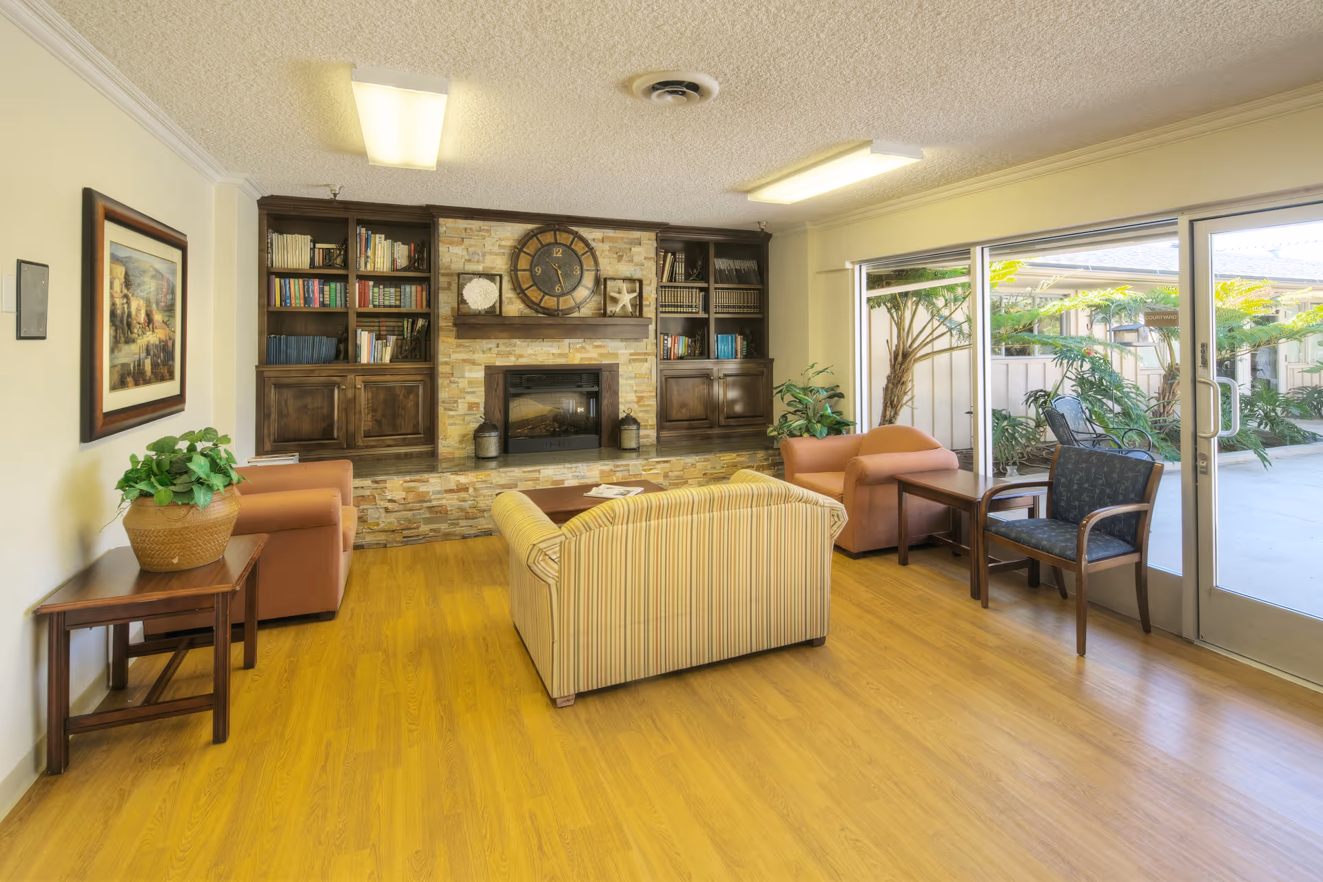 A cozy living room with wooden flooring, a striped sofa, two armchairs, a wooden side table with a potted plant, and a large built-in bookshelf surrounding a stone fireplace with a large clock above it. Large sliding glass doors open to an outdoor patio area with plants and chairs.