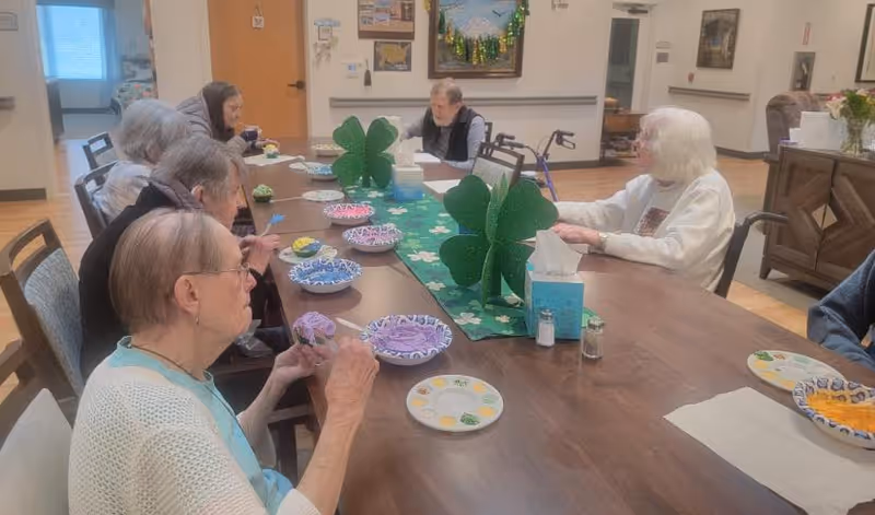 A group of elderly people sitting around a long wooden table in a communal room, engaging in an activity involving colorful bowls and plates. The table is decorated with green shamrock-shaped centerpieces and a green table runner with shamrock patterns. The room has light-colored walls, wooden flooring, and various decorations including a painting and bulletin board on the wall.