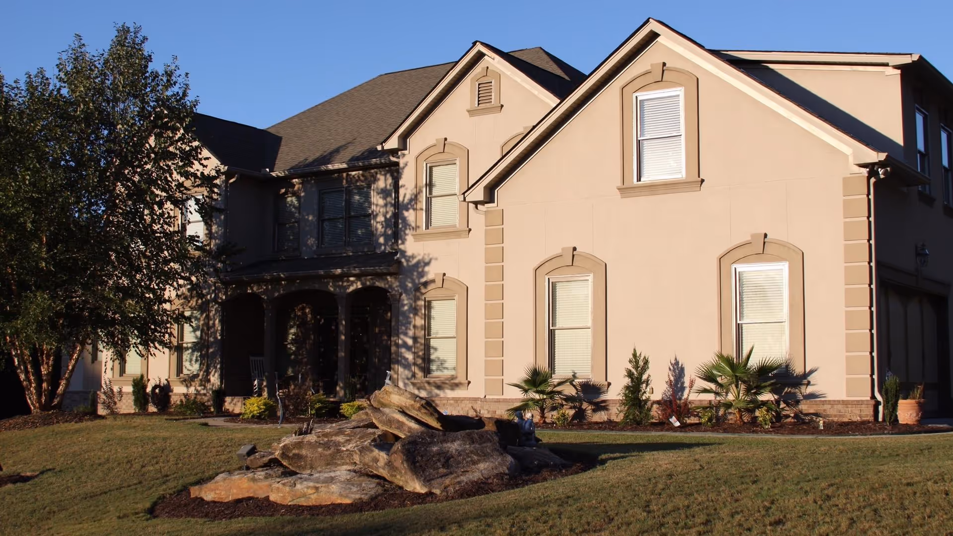 Front exterior of a two-story beige residential house with a landscaped lawn and rock feature in the yard.