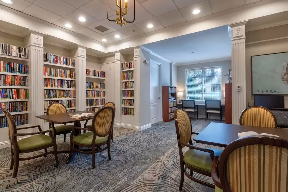 A cozy library room with multiple bookshelves filled with books along the walls. There are two tables with four wooden chairs each, featuring green cushioned seats and striped backs. The room has a patterned carpet, recessed ceiling lights, and a window with blinds letting in natural light. A framed map labeled 'Tampa' is visible on the wall.