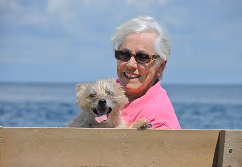 An elderly woman wearing sunglasses and a pink jacket is smiling while sitting on a bench by the water, holding a small fluffy dog that is panting with its tongue out.