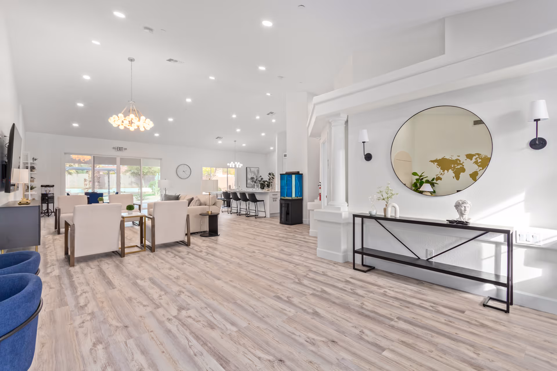 Bright and spacious senior living common area with light wood flooring, white walls, and modern furnishings. The room features a seating area with beige armchairs and a sofa, a large flat-screen TV mounted on the wall, a dining area with bar stools at a kitchen counter, and a decorative console table with a round mirror and wall sconces. Large windows and glass doors provide natural light and a view of the outdoor space.