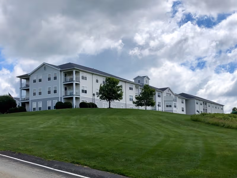 Three-story light-gray apartment-style building on a grassy hill under a cloudy sky.