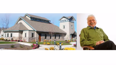 A senior living facility building with a stone and siding exterior, surrounded by landscaped flower beds and a clear blue sky. Next to the building image is a portrait of a smiling elderly man wearing a green sweater, sitting on a chair against a white background.