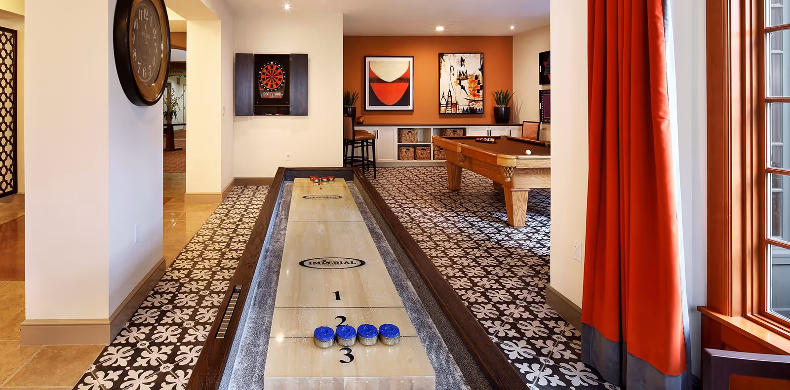 Interior recreation room with a shuffleboard table in the foreground, a pool table and dartboard in the background, patterned flooring and red curtains by a window.