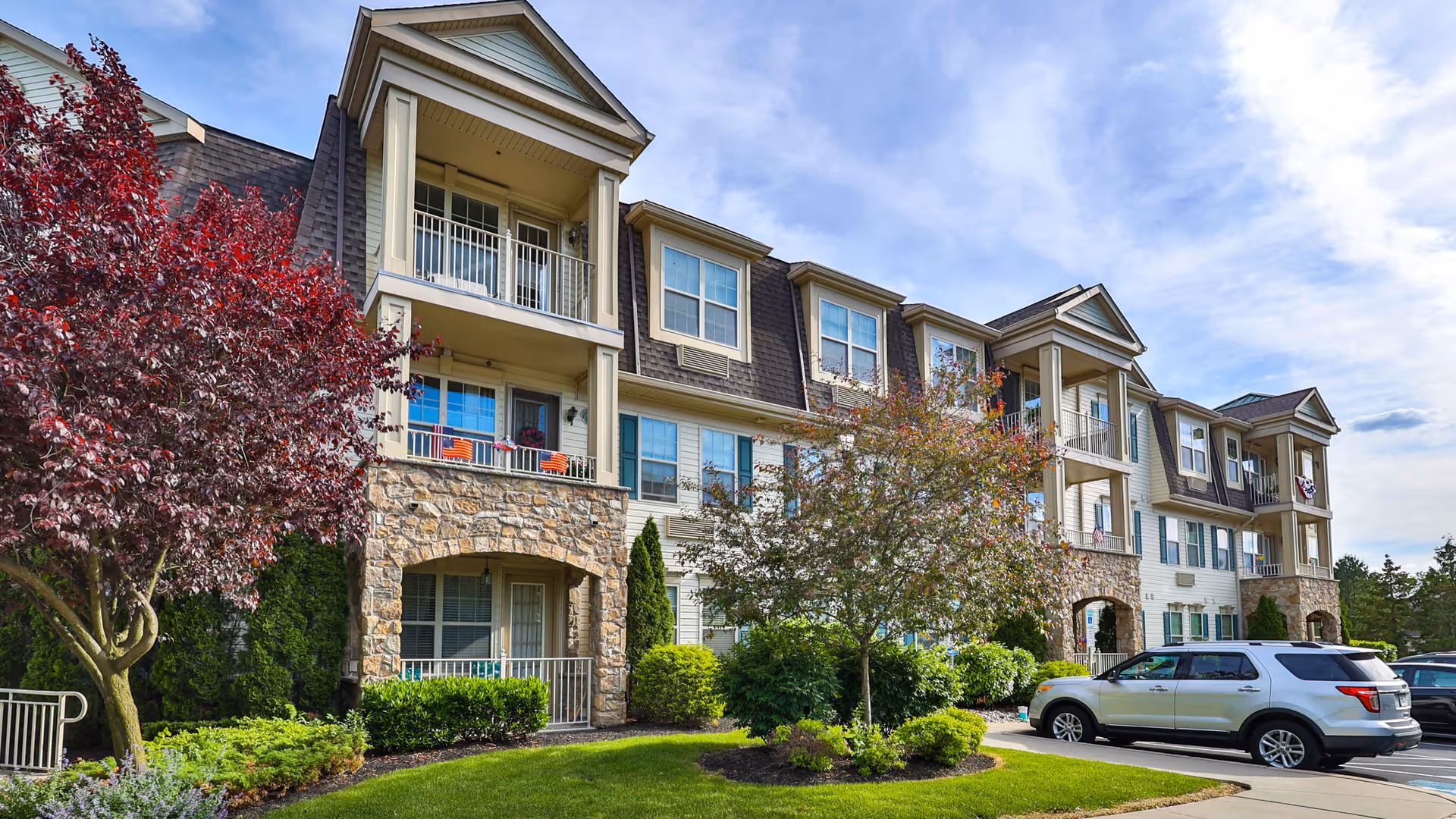 Three-story senior living building with balconies, stone accents, landscaped grounds, and parked cars.