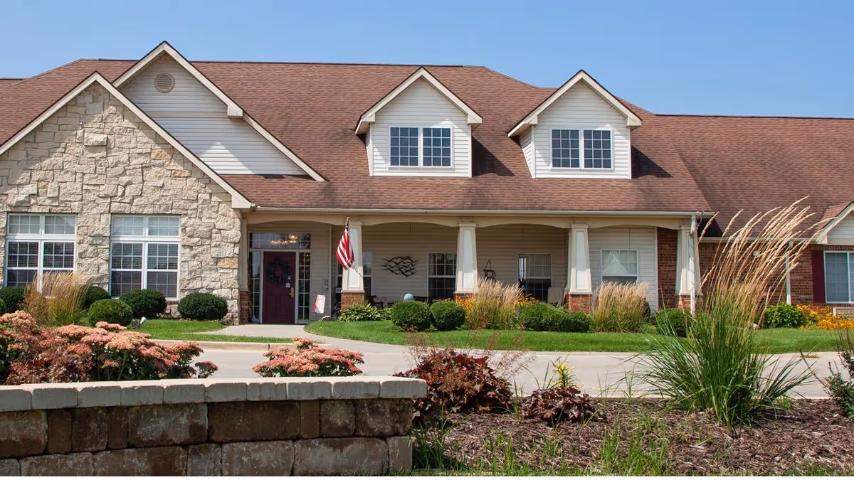 Front exterior of a single-story senior living building with a covered porch, gabled roof, American flag, and landscaped grounds.