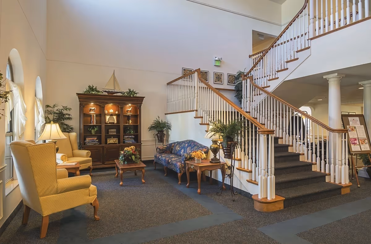 Bright lobby area with a grand staircase, upholstered seating, a wooden display cabinet, and decorative plants.
