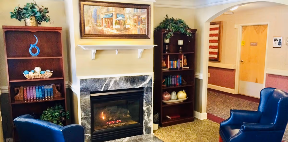 A cozy sitting area in Cambridge House featuring a fireplace with a marble surround, two blue leather armchairs, two wooden bookshelves filled with books and decorative items, a framed painting above the fireplace, and a hallway with a door in the background.