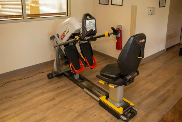 A recumbent exercise machine with foot pedals and a seat is placed on a wooden floor in a room with beige walls. A window with blinds is visible on the left side, and a red fire extinguisher is mounted on the wall in the background.