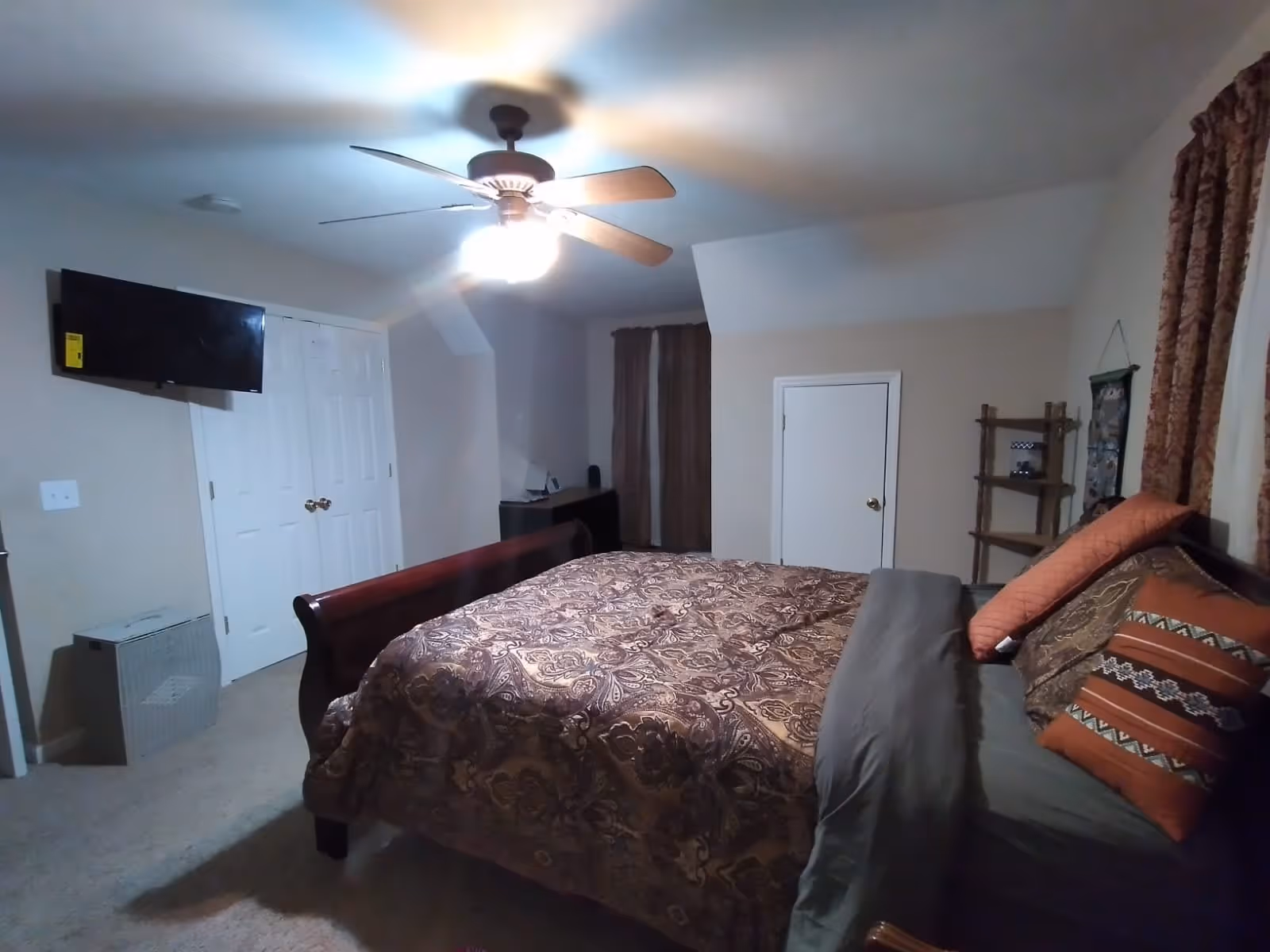 A bedroom with a large bed covered in a patterned brown and beige comforter and multiple pillows. The room has beige walls, a ceiling fan with lights, a wall-mounted TV, a double-door closet, a small desk with items on it, and a wooden shelf unit. There are brown curtains on the windows.