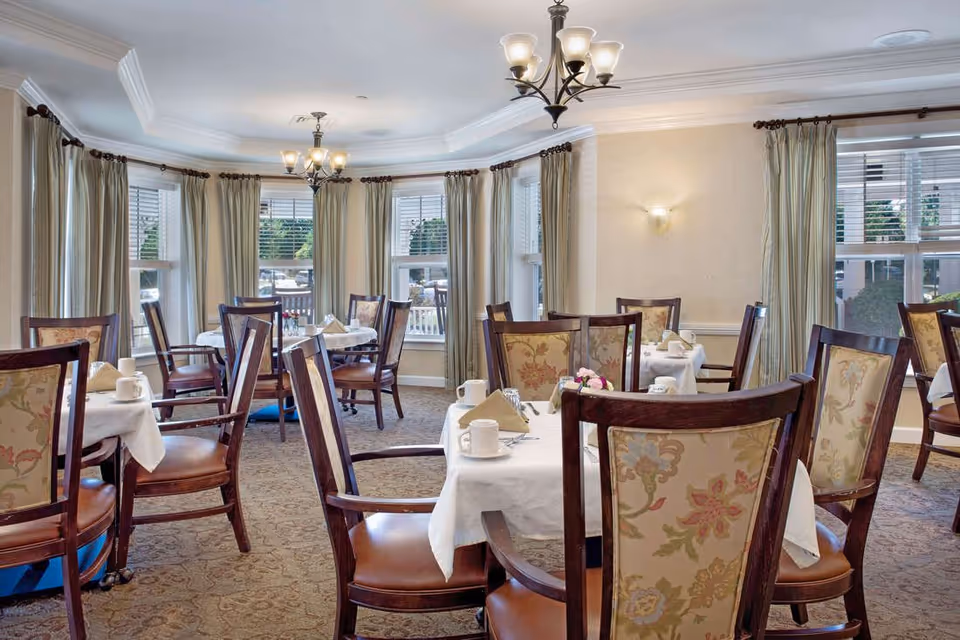 Bright dining room with round tables set for a meal and upholstered wooden chairs in front of bay windows.