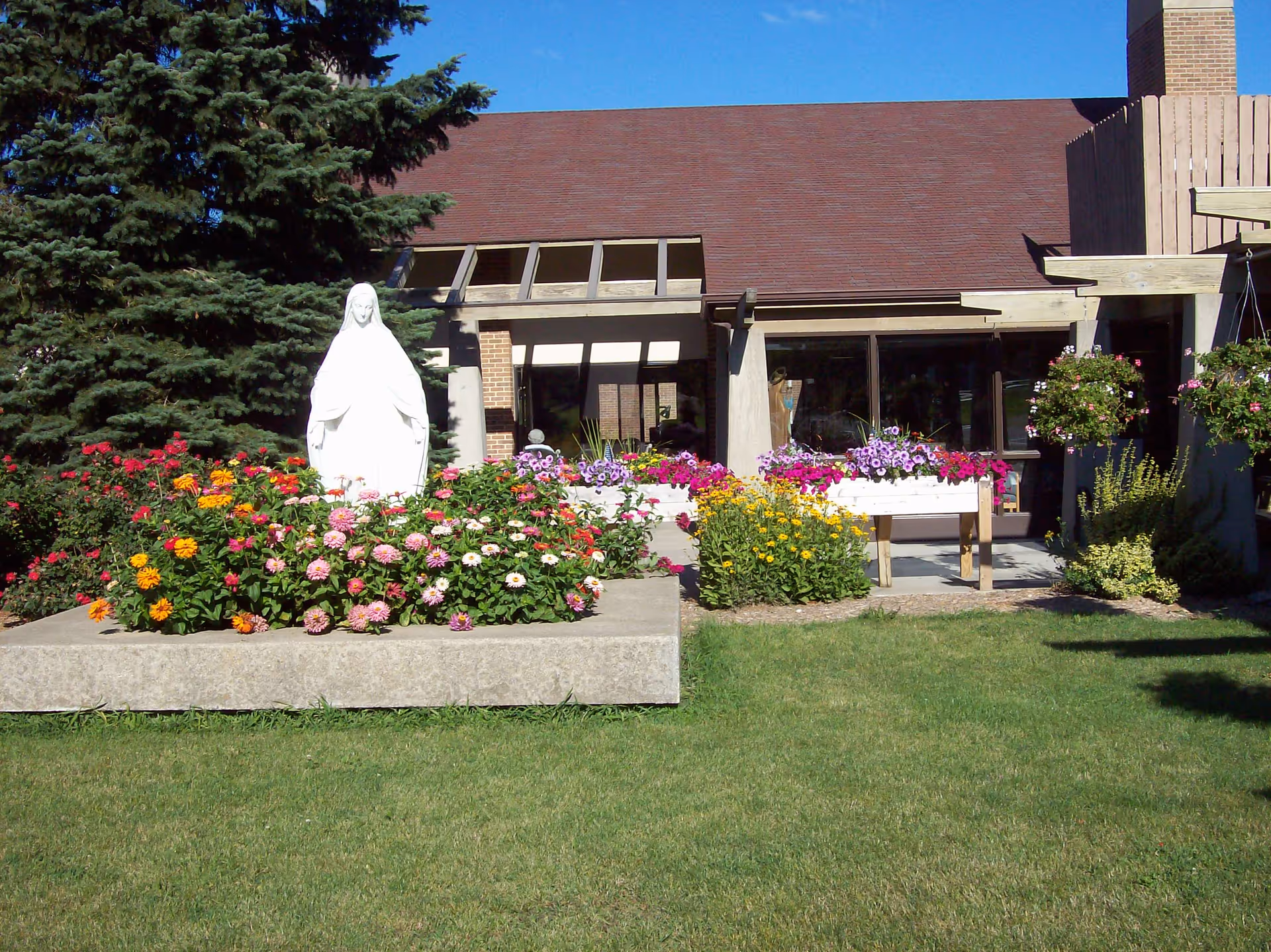 Outdoor garden area at St Monica's Senior Living featuring a white statue of a robed figure surrounded by colorful flowers and greenery, with a building and large windows in the background under a clear blue sky.
