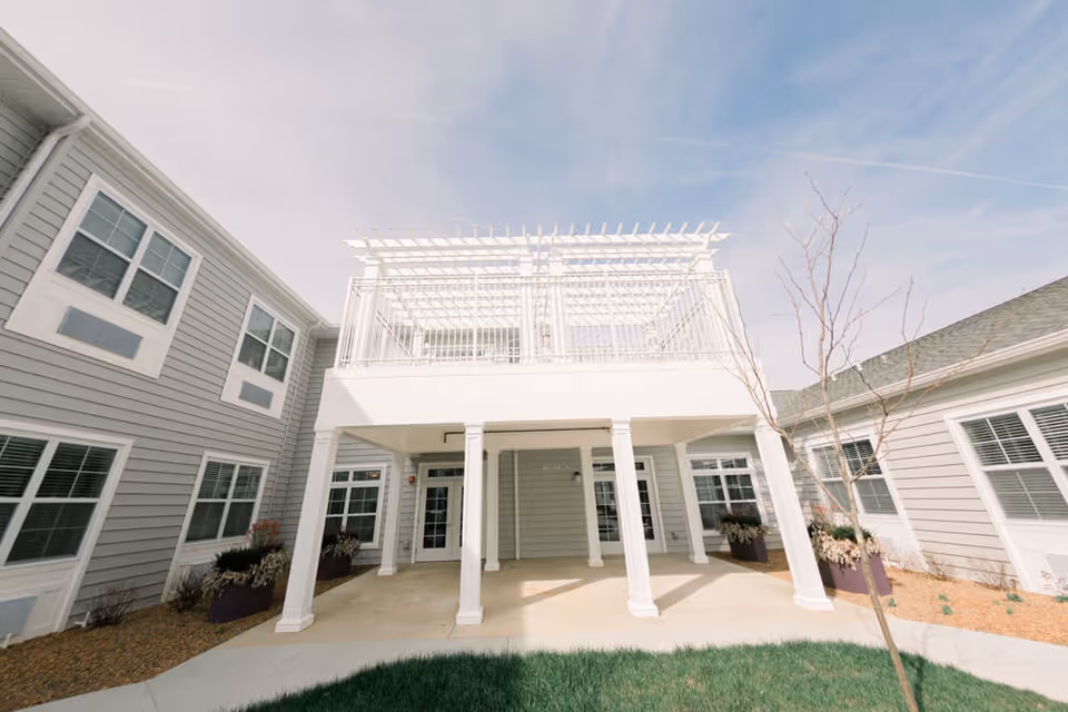 Exterior view of a senior living facility building with gray siding, white trim, and multiple windows. The building features a covered patio area supported by white columns and an upper balcony with a white pergola. There are some small trees and planters with plants around the patio area under a partly cloudy sky.