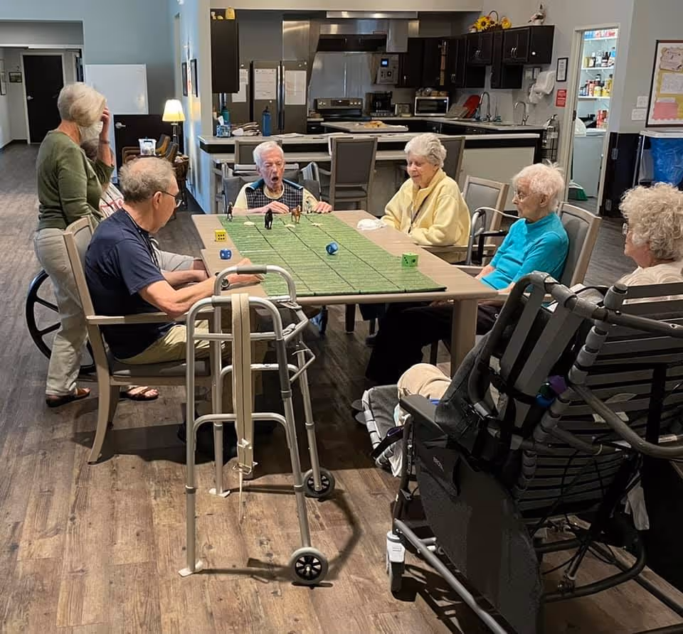 A group of elderly people sitting around a table in a common area, playing a game with dice and small horse figurines. The room has a kitchen in the background with dark cabinets and stainless steel appliances. Some mobility aids like a walker and a wheelchair are visible.
