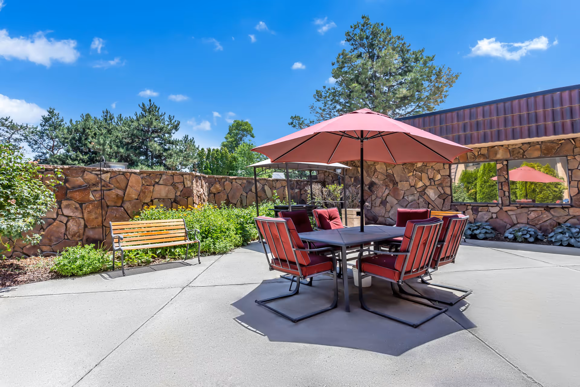 Outdoor patio area with a square table surrounded by six red cushioned chairs and a large red umbrella providing shade. There is a wooden bench to the left, stone walls surrounding the patio, and trees and plants in the background under a clear blue sky.