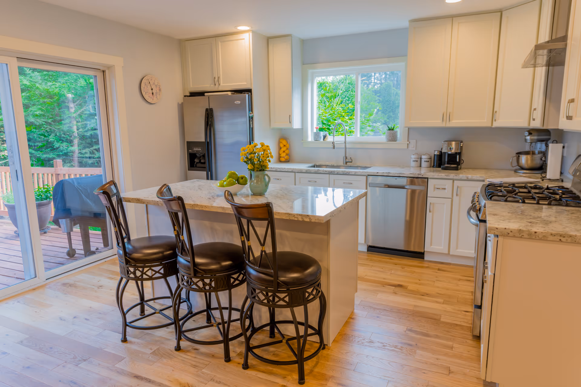 Bright kitchen with white cabinets, stainless steel appliances including a refrigerator, dishwasher, and gas stove. There is a kitchen island with a marble countertop and three high chairs. A window above the sink shows greenery outside, and sliding glass doors lead to a wooden deck with a grill.